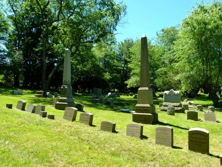 Historic Greenville Cemetery with old headstones and two obelisks on a sunny lawn.